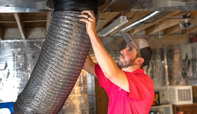 A technician connecting a large, flexible vacuum hose to overhead metal ductwork for air duct cleaning.