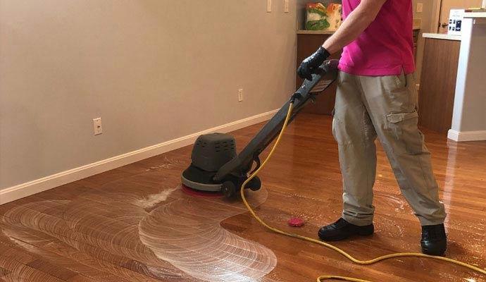 Professional floor technician in a pink shirt using a power buffer to remove dull buildup and clean a hardwood floor.